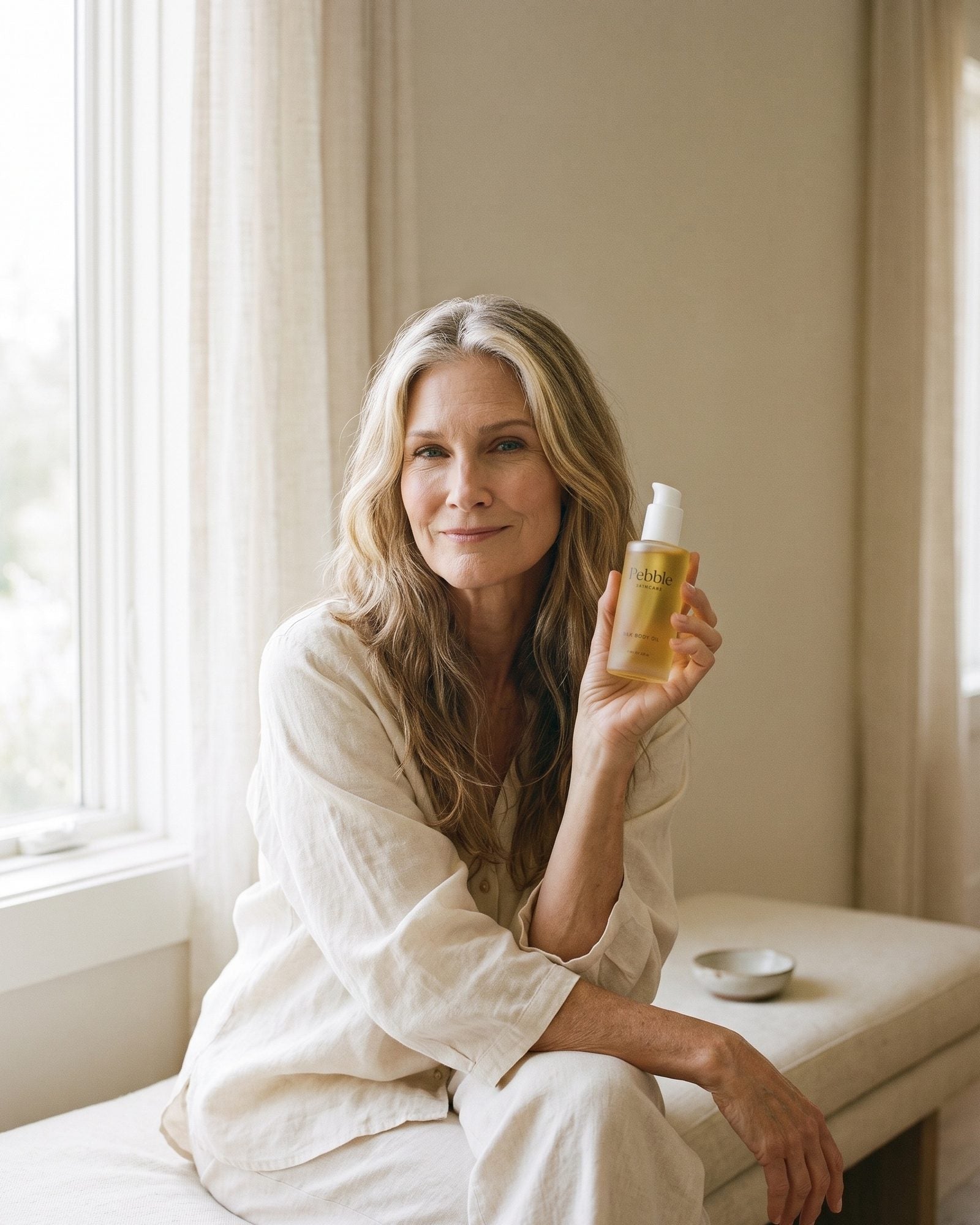 Woman in her fifties holding Pebble Silk Body Oil, soft natural light on glowing arm skin
