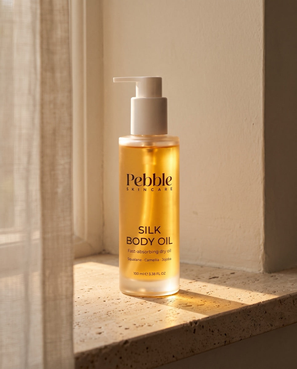Pebble Silk Body Oil glowing in warm window light on a travertine sill